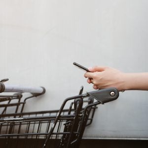 close up asian woman hand using smart phone and holding trolley bar for shopping on cement background at outside mall concept.