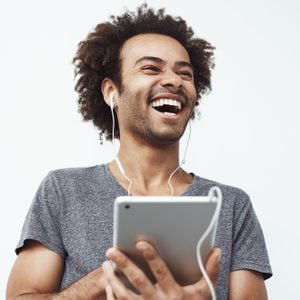 Portrait of cheerful happy african man in headphones laughing holding tablet talking or watching and enjoying a comedy show or browsing over white background.