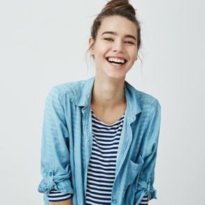 Girl likes funny jokes. Smart good-looking student with bun hairstyle trembling from laugh, smiling positively and being in great mood while standing over gray background. Woman watch hilarious show