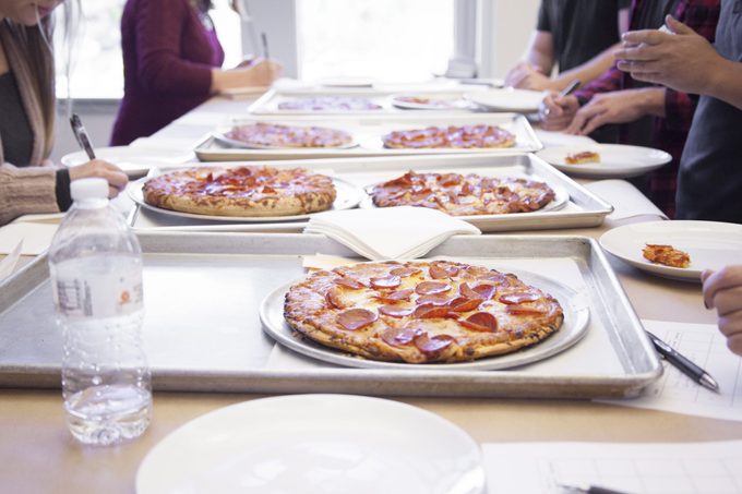 Table filled with unmarked pizzas with taste testers gathered around