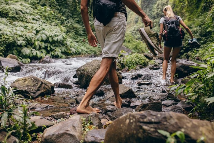 Low section shot of man and woman crossing the stream barefooted. Couple walking by the creek in forest with their shoes in hand. Hikers hiking in forest.