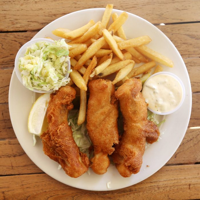 fish and chips with French fries and pineapple coleslaw on a white plate on a wooden table.