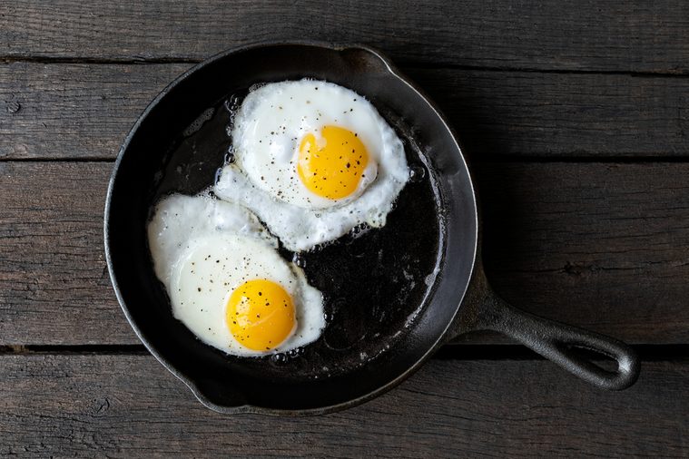 Two fried eggs in cast iron frying pan sprinkled with ground black pepper. Isolated on dark painted wood from above.