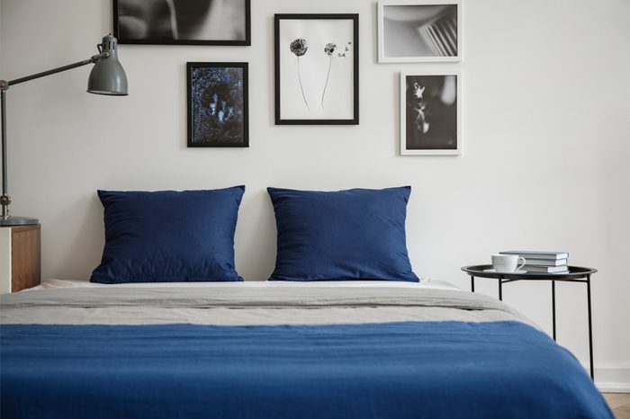 Navy blue pillows on bed between table and lamp in white bedroom interior with posters. Real photo