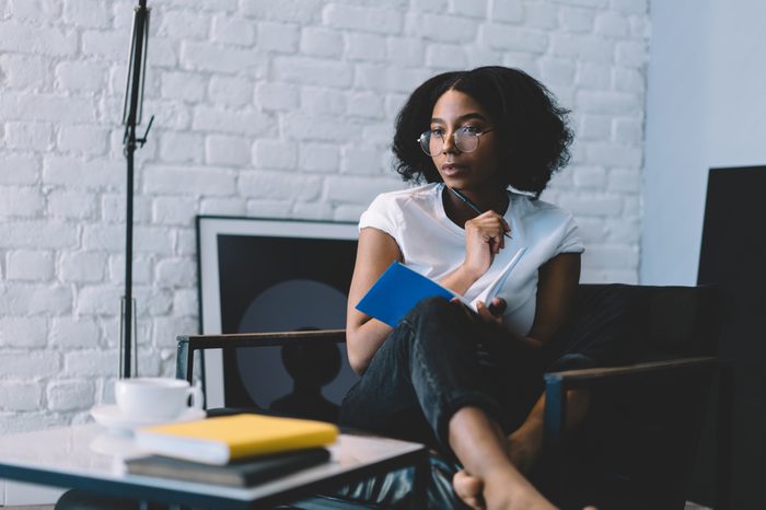 Thoughtful african american young woman in optical eyeglasses holding pen and thinking on creative ideas for writing her blog publication in notebook enjoying leisure time at home interior apartment