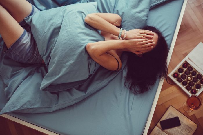 Depressed woman in bed with hands over face. box of chocolates and a glass of wine on the floor nearby.