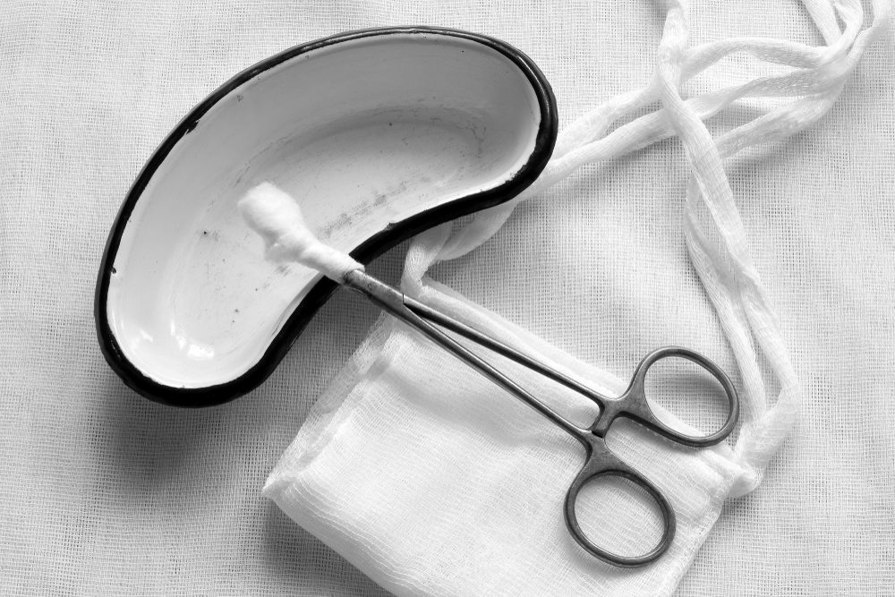 Retro. Gauze medical mask, metallic forceps with a cotton swab and a kidney-shaped enamelled metal patch on a white gauze background. Black and white photography.