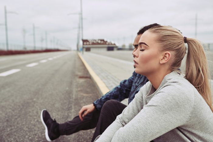 Diverse young couple in sportswear sitting on the side of a road together taking a break from a run on an overcast day