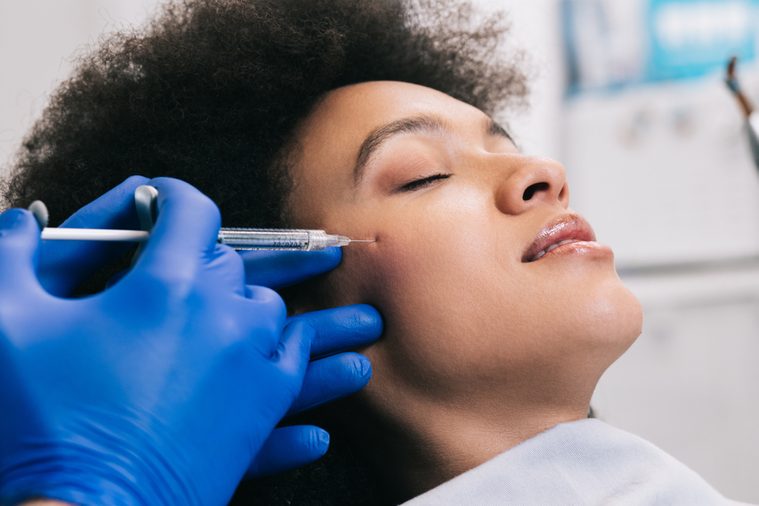 Attractive young African woman is getting a rejuvenating facial injections. She is sitting calmly at clinic. The expert beautician is filling female wrinkles by hyaluronic acid.