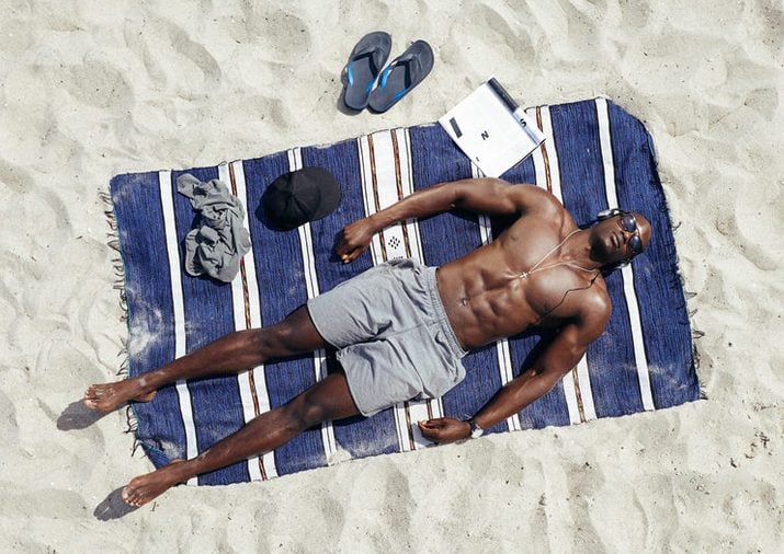 Top view of young guy lying shirtless on a mat sunbathing. African male model relaxing on beach on a hot summer day.