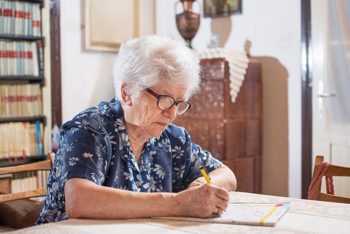 Elderly woman with eyeglasses sitting near the table and doing crossword.