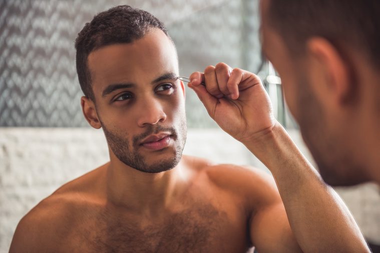 Handsome naked Afro American man is plucking eyebrows while looking into the mirror in bathroom
