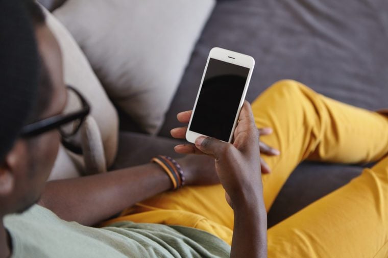 Unrecognizable stylish Afro American man wearing yellow jeans relaxing at home, lying on comfortable sofa, reading message on smart phone with black blank copy space screen for your information