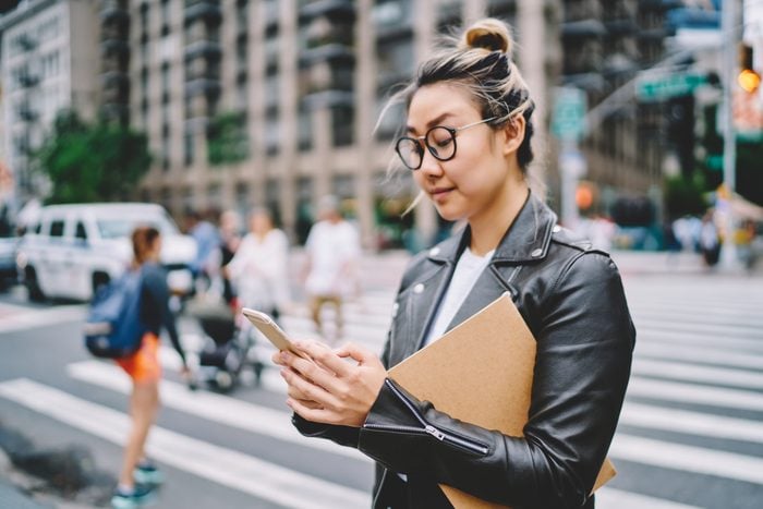 Young attractive female blogger in glasses for eye protection sharing news with followers via smartphone application.Gorgeous hipster girl strolling at urban setting with gadget during leisure time