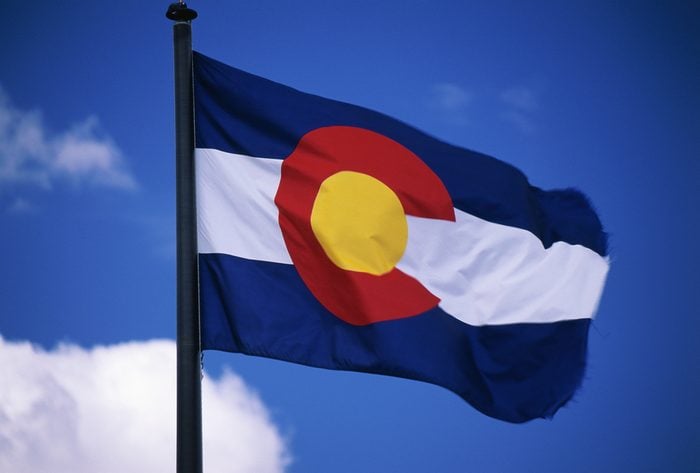 This is the Colorado State Flag, waving in the wind situated on a flag pole. It is set against a blue sky. At the center of the flag is a large capital C.