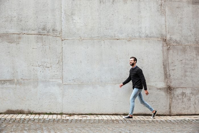 Full length portrait of a smiling casual man walking on a city street against gray wall