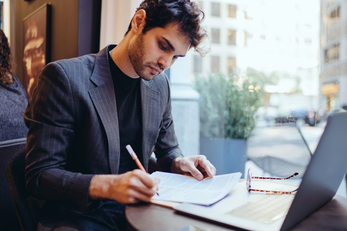 Young handsome caucasian businessman  in trendy suit writing information on financial papers for banking on laptop computer, serious confident male doing paperwork remotely indoors in coffee shop 