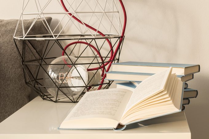 White nightstand with wire lampshade and large incandescent bulb standing next to a pile of books in blue cover, one of which is lying open