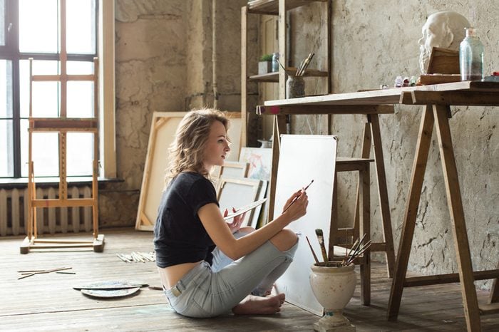 Woman painter sitting on the floor in front of the canvas and drawing. Artist studio interior. Drawing supplies, oil paints, artist brushes, canvas, frame. Workshop or art class. Creative concept