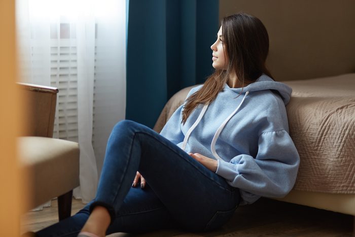 Indoor shot of thoughtful dark haired woman dressed in blue sweatshirt, jeans, sits on floor near bed, looks thoughtfully at window, poses in cozy bedroom at modern apartment. Domestic atmosphere
