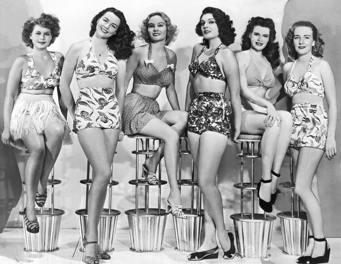VARIOUS United States: c. 1952. Six attractive young women in two piece bathing suits sit in a row on modern stools.