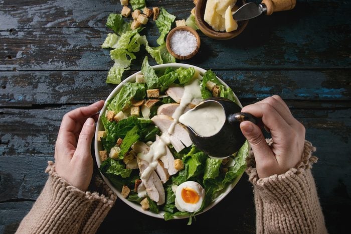 Female hands powring dressing to Classic Caesar salad with chicken breast in white ceramic plate. Served with ingredients above over old dark blue wooden background. Flat lay. Rustic style