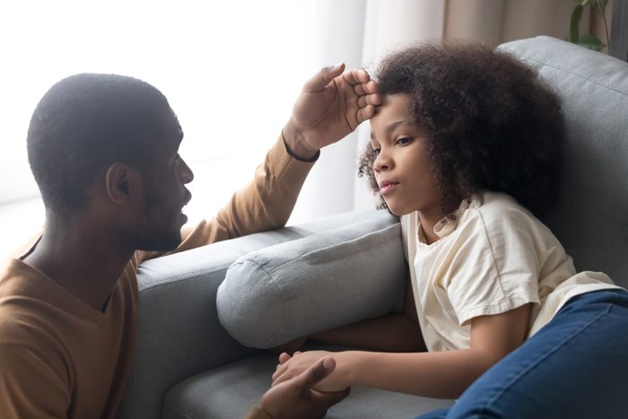 Caring african father touching forehead of sick ill kid daughter lying on couch, worried black dad checking child temperature, fever flu symptoms and treatment, children medical health care concept