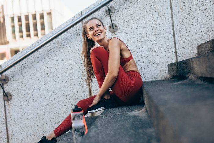 Smiling woman in sportswear sitting on steps outdoors after a run. Female athlete taking break after running workout.