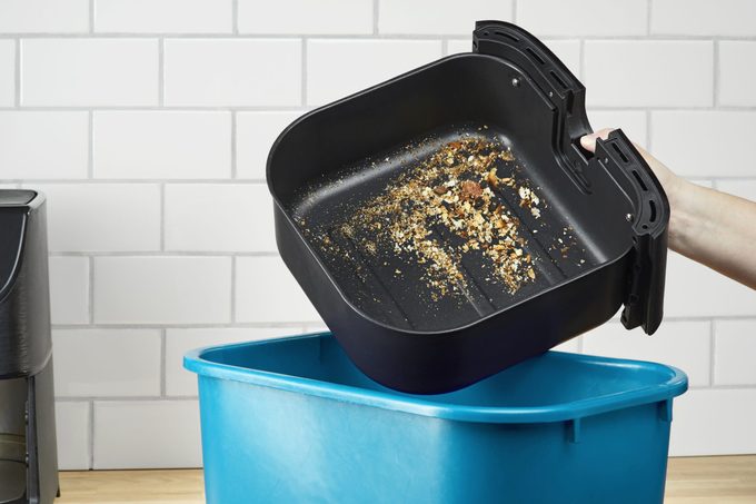 pouring the crumbs from an air fryer into a trash can in a kitchen