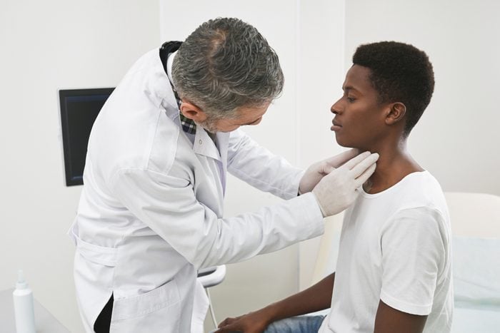 Therapist examining neck of african patient in medical cabinet. Doctor palpating lymph node with fingers. Physician wearing in white medical gown and gloves. Man wearing in white t shirt.