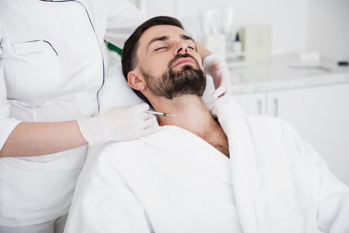 Man in white bathrobe staying calm while experienced cosmetologist in rubber gloves carefully touching his neck and making an injection
