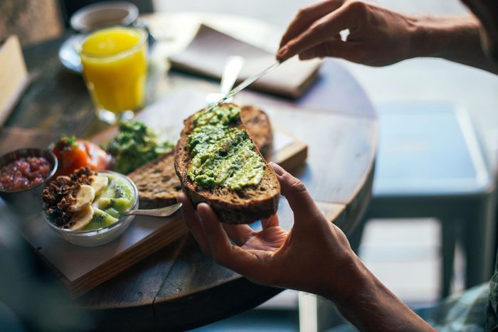 Soft focus shot of man having delicious huge breakfast at cool restaurant or cafe, puts guacamole or avocado spread on top of rye bread toast, ready to indulge and fullfill hunger