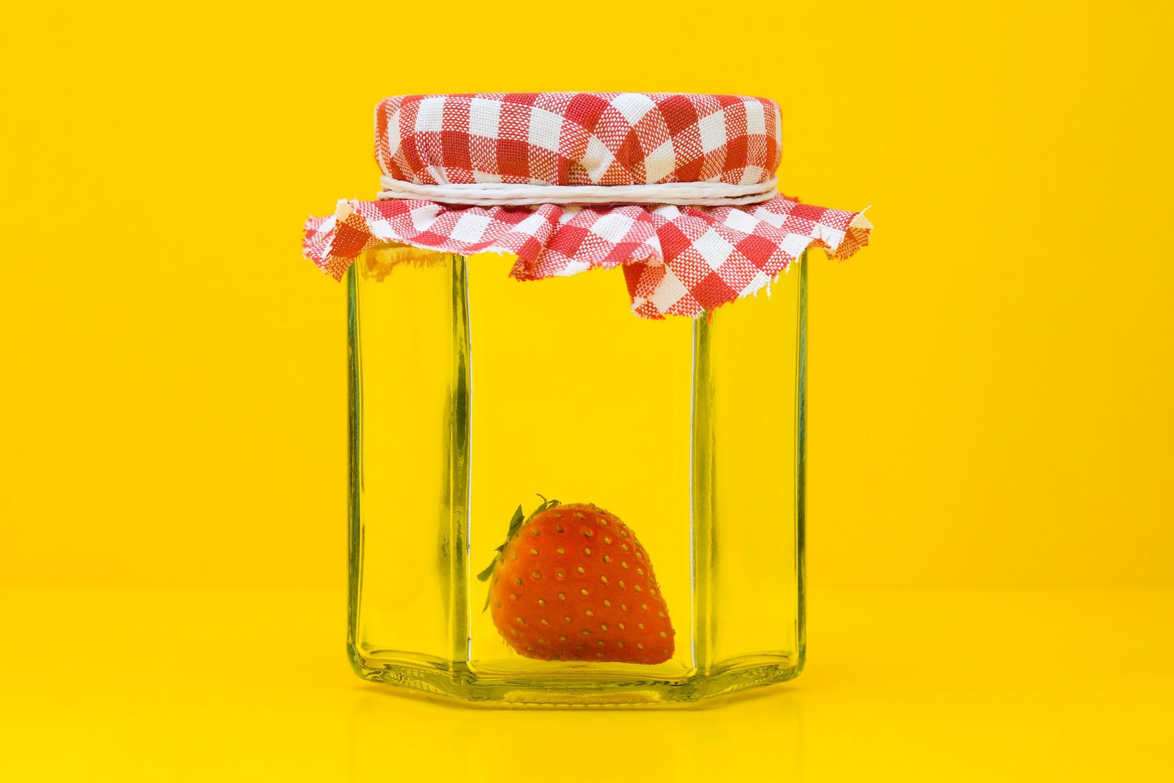 a single fresh strawberry inside a glass jar used for jam or jelly on a yellow background