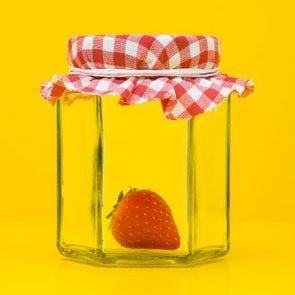 a single fresh strawberry inside a glass jar used for jam or jelly on a yellow background