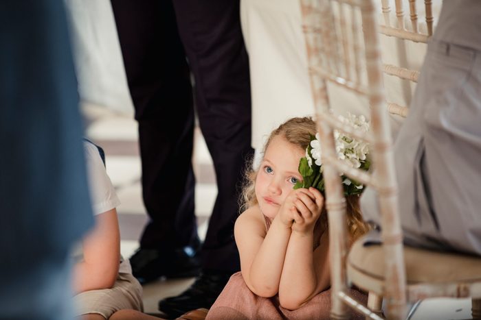 Little girl is sitting on the dancefloor by a table at a wedding. She is watching the bride and groom share their first dance. 