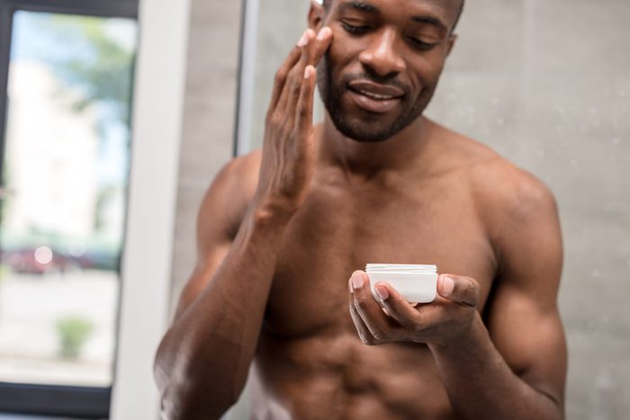 smiling shirtless young african american man applying facial cream in bathroom