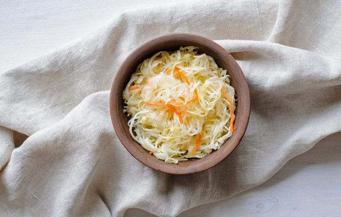 Top view of brown ceramic bowl of sauerkraut with chopped cabbage and carrot on grey towel sitting on white table surface