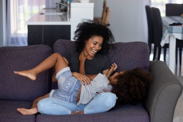 Happy african american mom tickling kid daughter laughing together on couch, cheerful black mother playing funny game with little cute girl at home, mixed race child having fun relaxing with mum