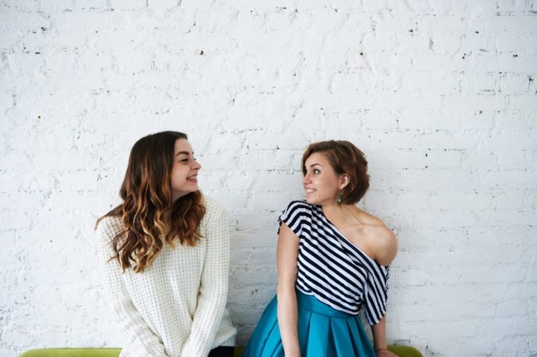 People, youth, happiness, joy and friendship. Two happy beautiful European young women friends or siblings wearing trendy clothes, posing at white brick wall, looking at each other and smiling