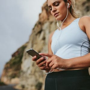 Young female runner taking a break and listening to music during the run outdoors. Young woman with headphones looking at mobile phone.
