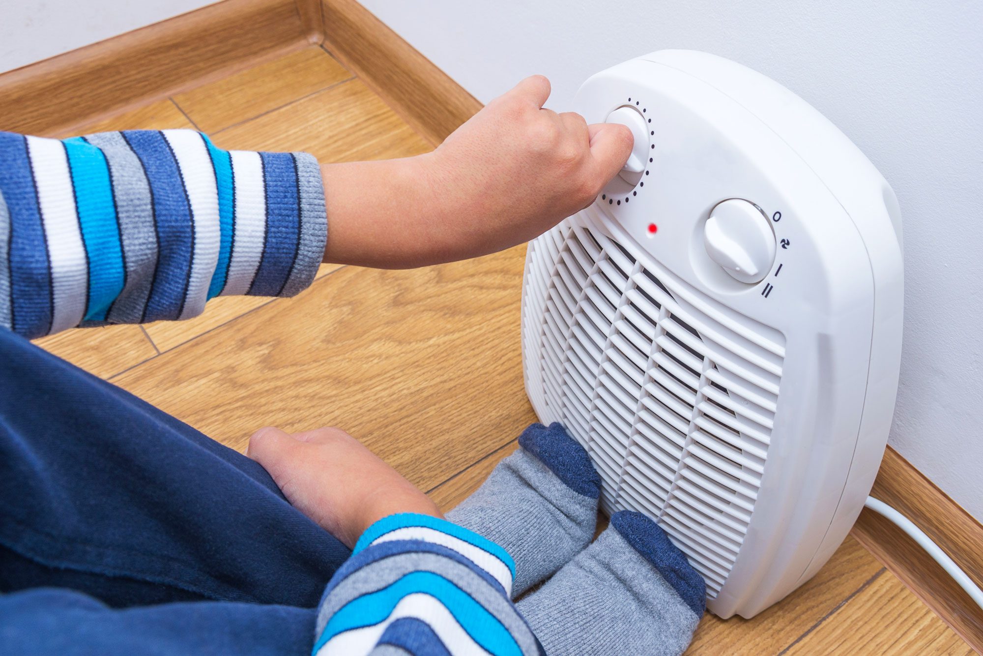 A Young Boy Warms Himself Near An Electric Fan Heater Sitting On The Floor