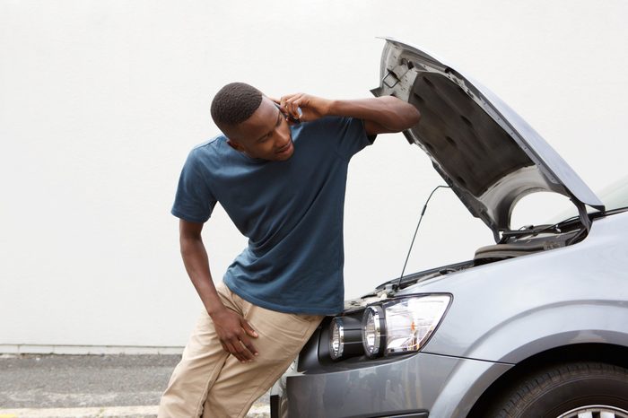 Portrait of young african guy standing near broken car and calling for help on mobile phone