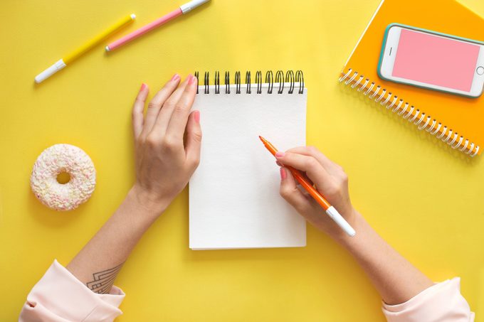 Colorful yellow background with stationary, donut, smart phone mock-up woman's hands writing in empty notebook. Flat lay top view. Learning to draw, making wish list or plans. Art education concept