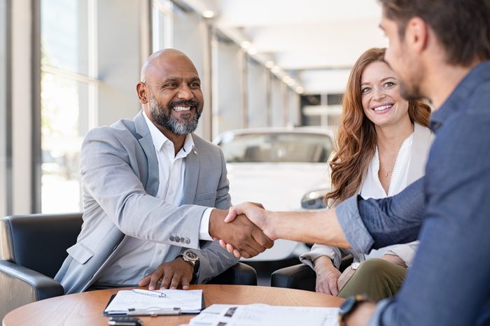 Smiling couple buying a new auto at car showroom. African salesman making handshake with young man purchasing new car with his girlfriend. Happy african agent shaking hands with customer couple.