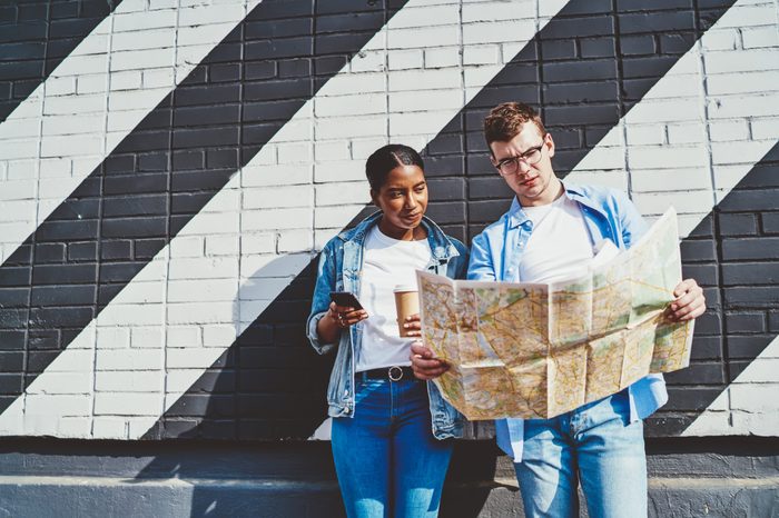 Caucasian young man together with African American hipster girl reading travel map to search right route during trip standing near copy space wall. Diverse couple millennial of tourists sightseeing 