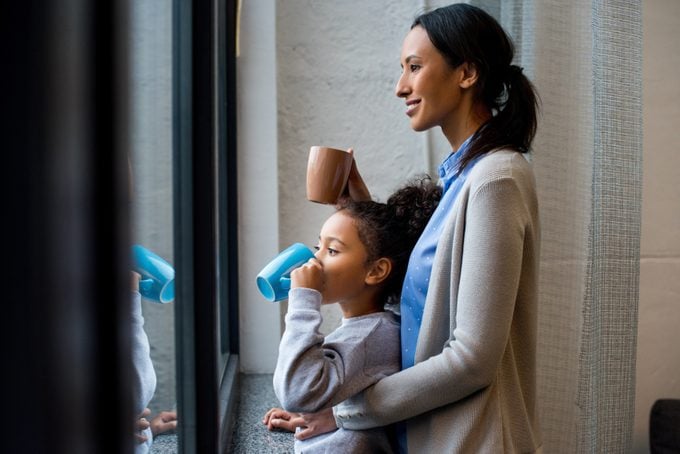 african american mother and daughter drinking hot beverage at window