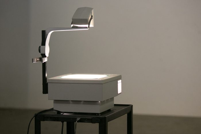 A vintage overhead projector sit on a roller cart lighting a wall ready to show overhead projection transparencies. Overhead projectors were often used in school & business before digital projection.