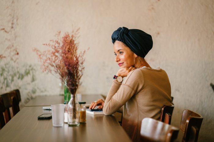 Portrait of a young Muslim woman. She is of Asian descent (Arab, Malay, Asian) and is wearing a turban (hijab, head scarf) and is smiling as she sits at a wooden table with flowers with her devices.
