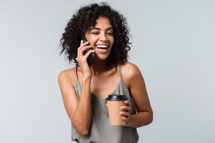 Happy young african woman casually dressed standing isolated over gray background, talking on mobile phone, holding takeaway cup of coffee