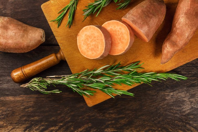 An overhead photo of slices of sweet potatoes with branches of fresh rosemary, shot from above on dark wooden textures with a place for text
